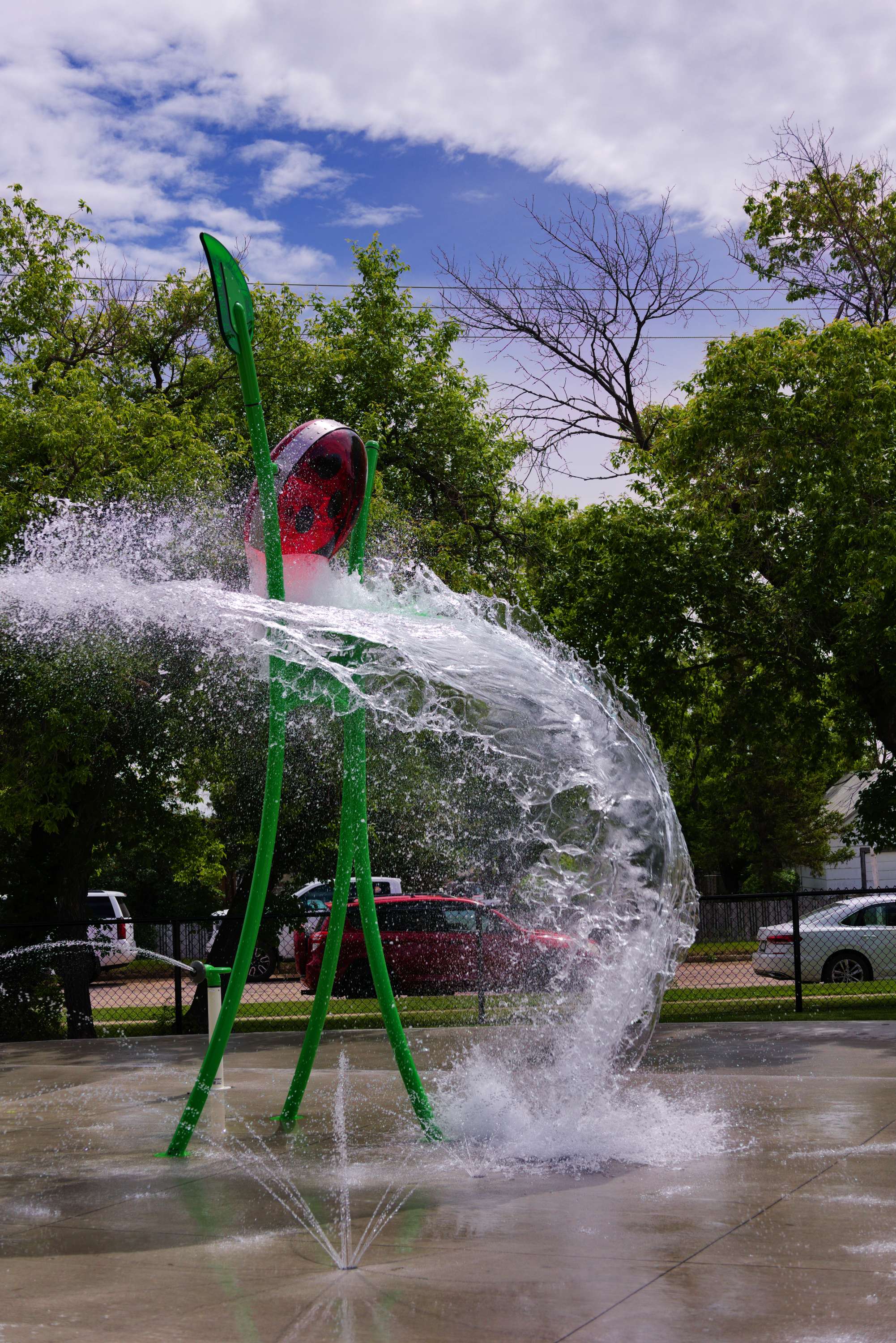 Ladybug bucket dumping water at a spray park with water jets shooting upward on a sunny day.