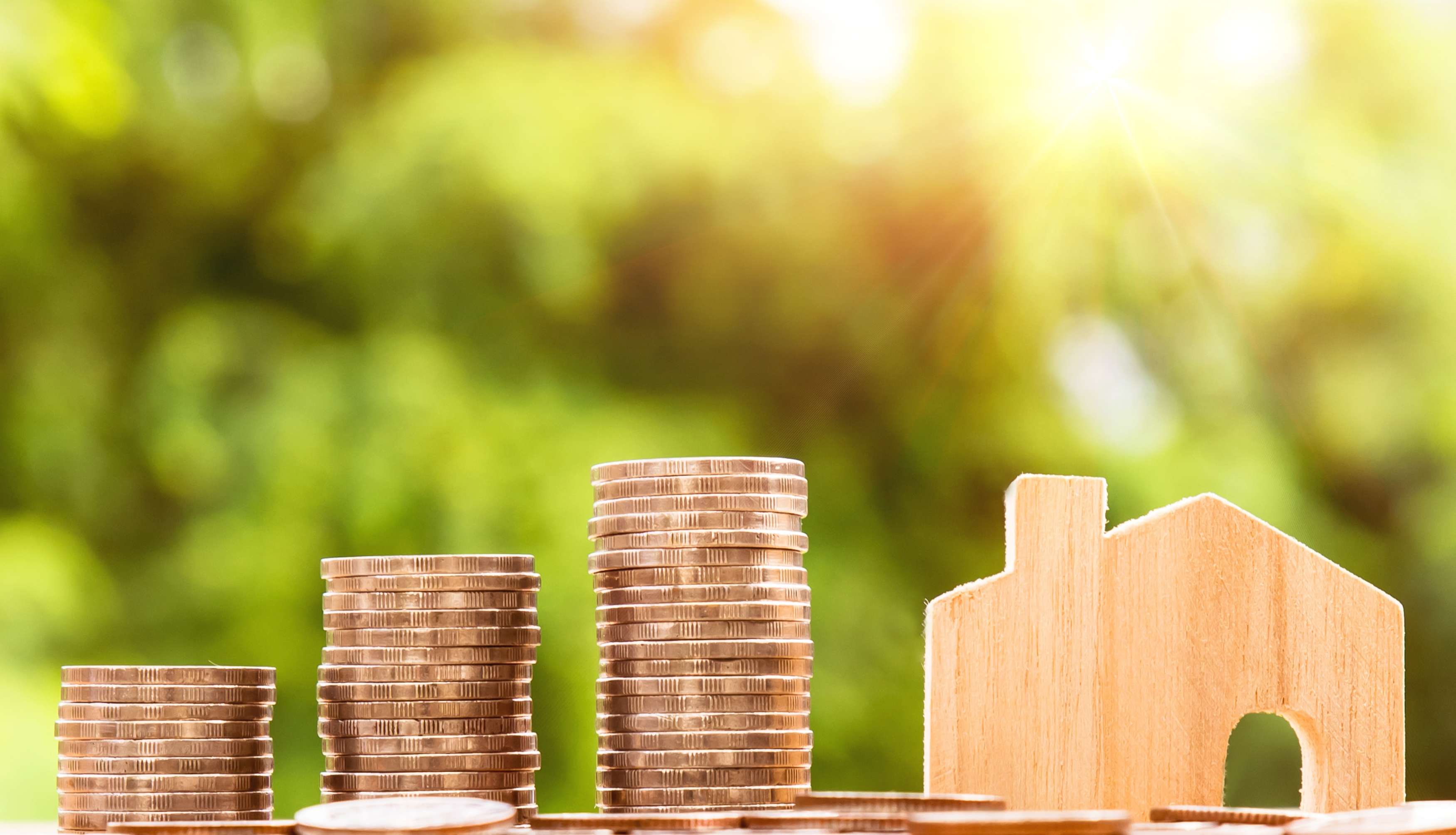 Stacks of coins beside a small wooden house model with sunlight in the background.