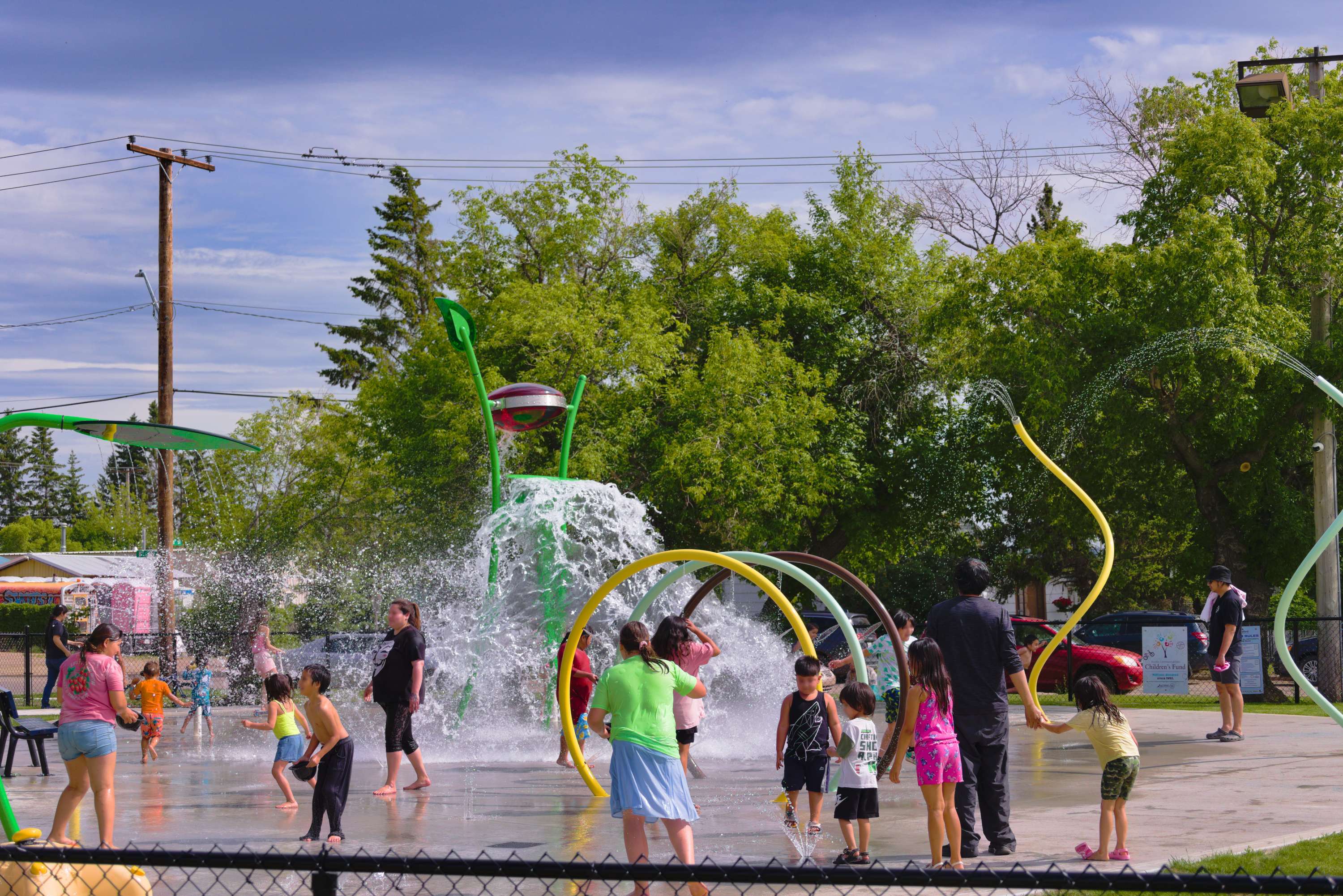 People playing under a spray park water fountain with jets rising and surrounding trees.
