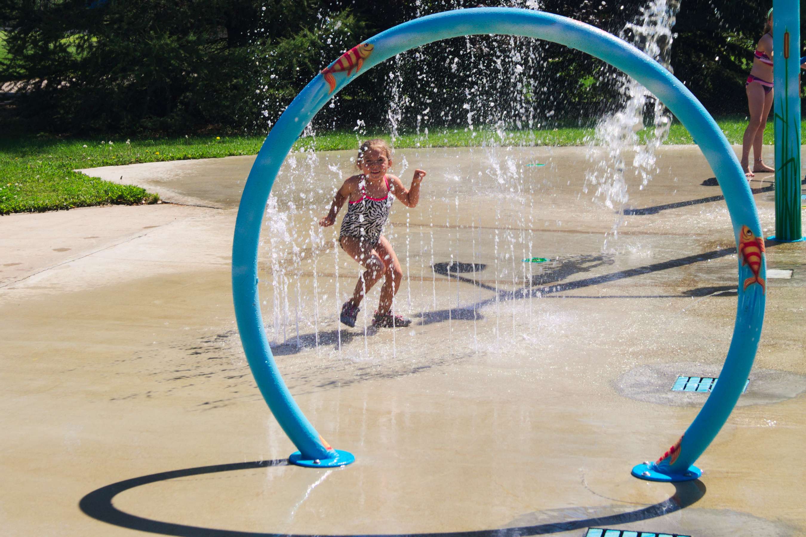 Child running through a circular spray feature at a sunny outdoor splash park.