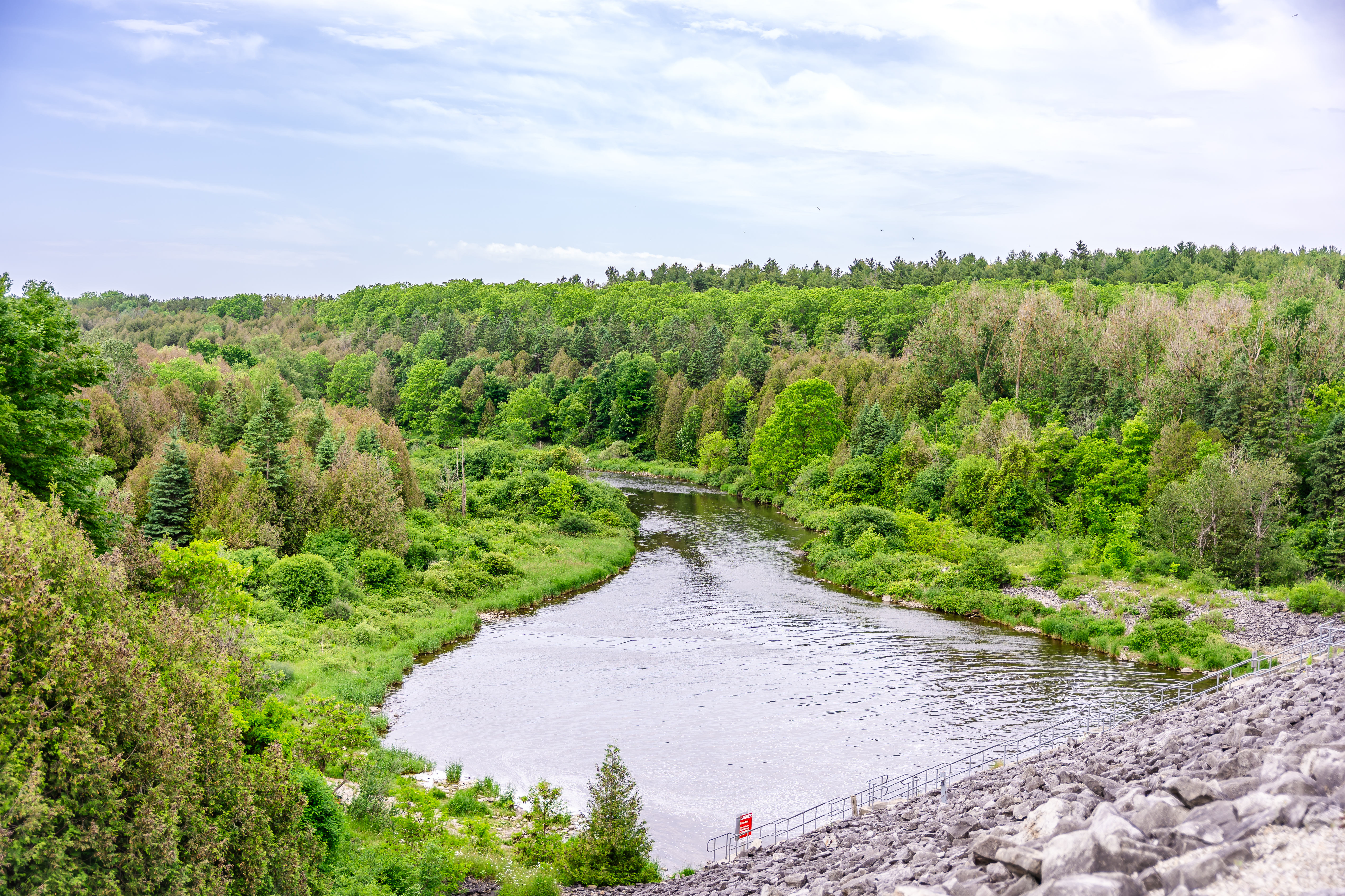 View of a winding river bordered by dense green forest, seen from an elevated vantage point. A rocky embankment slopes down on the right side toward the water under a partly cloudy sky.