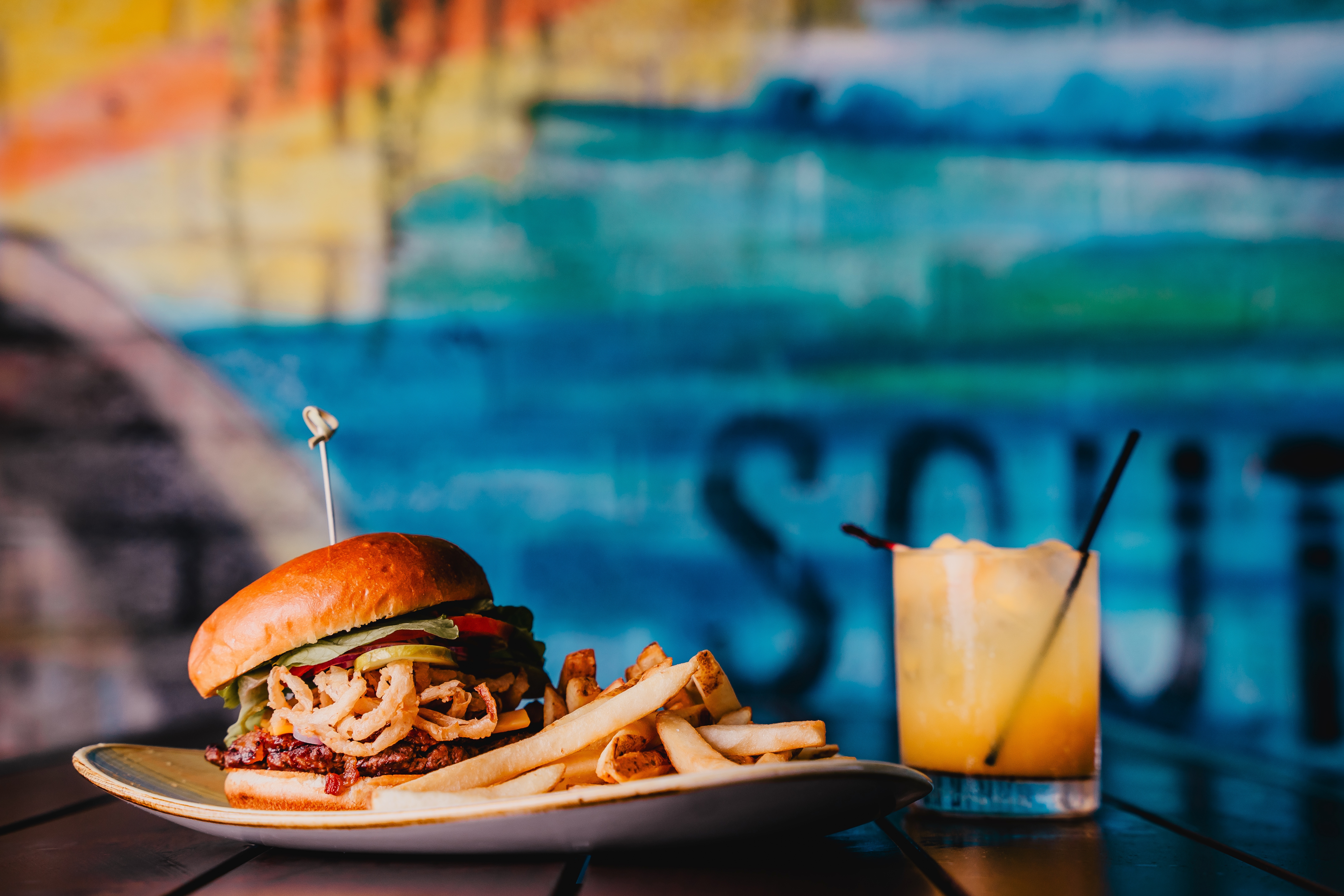 A burger and fries on a plate alongside a colourful drink sit in front of a blue background
