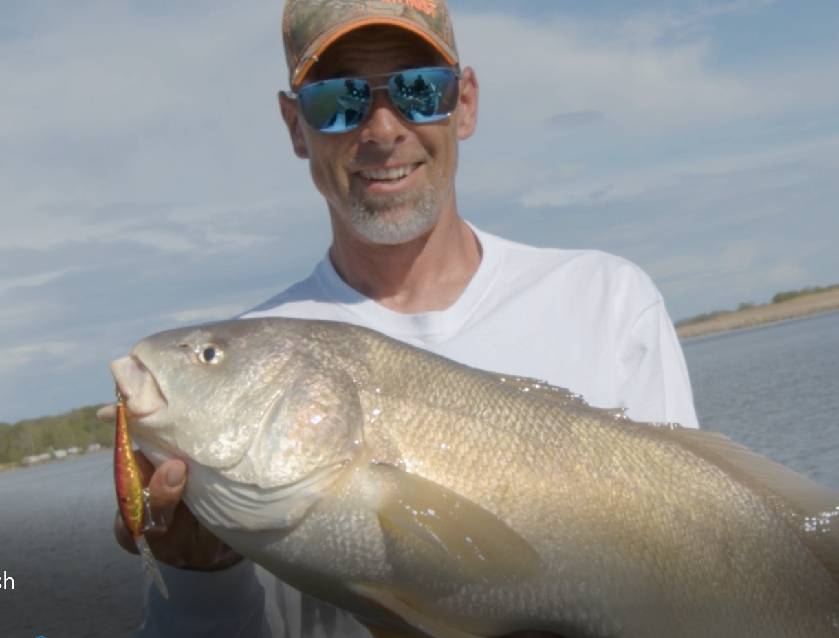A man smiles holding a large beige fish with the Grand River in the background