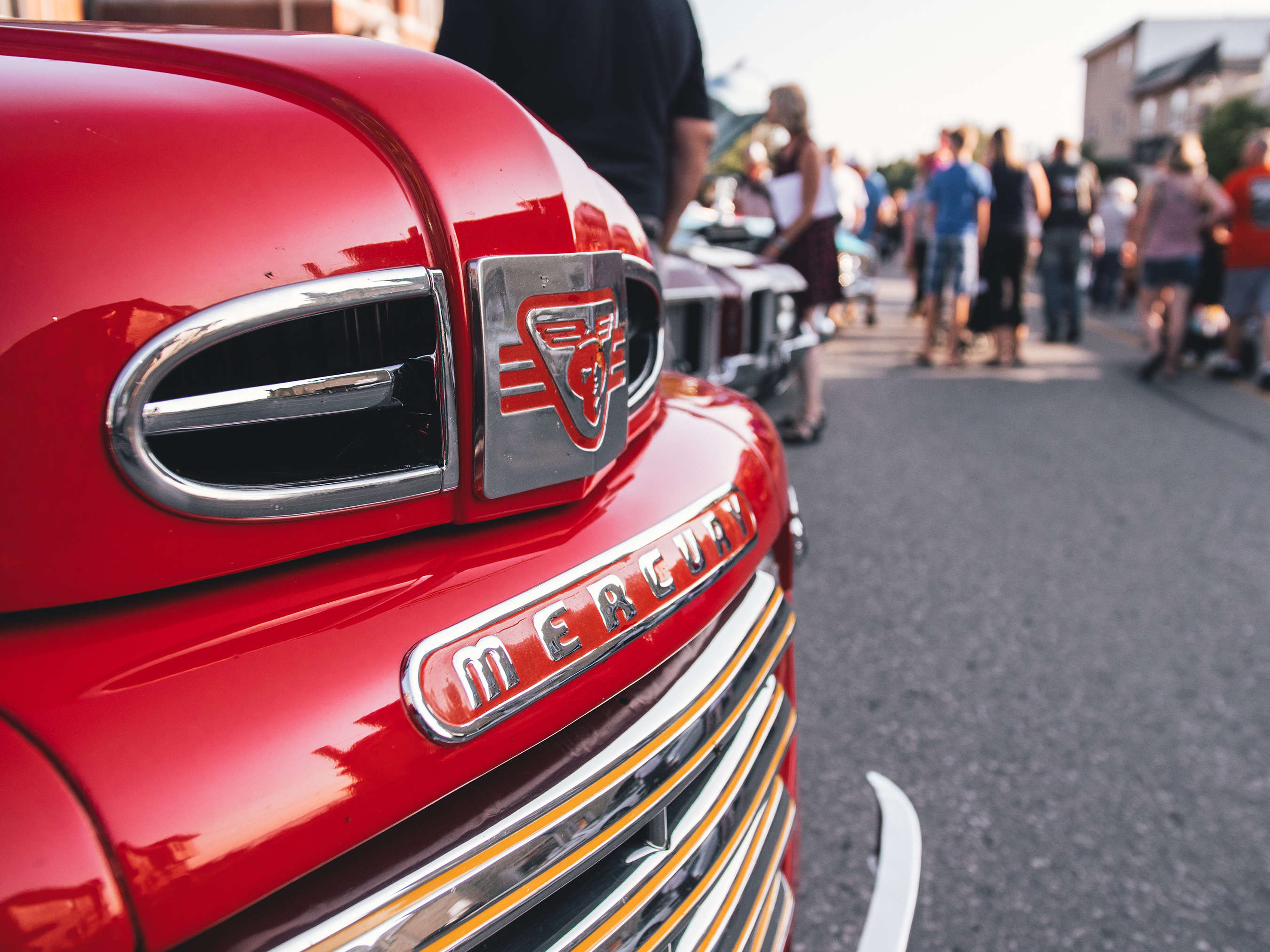 The front grill of a red classic car with crowds of people standing just beyond.