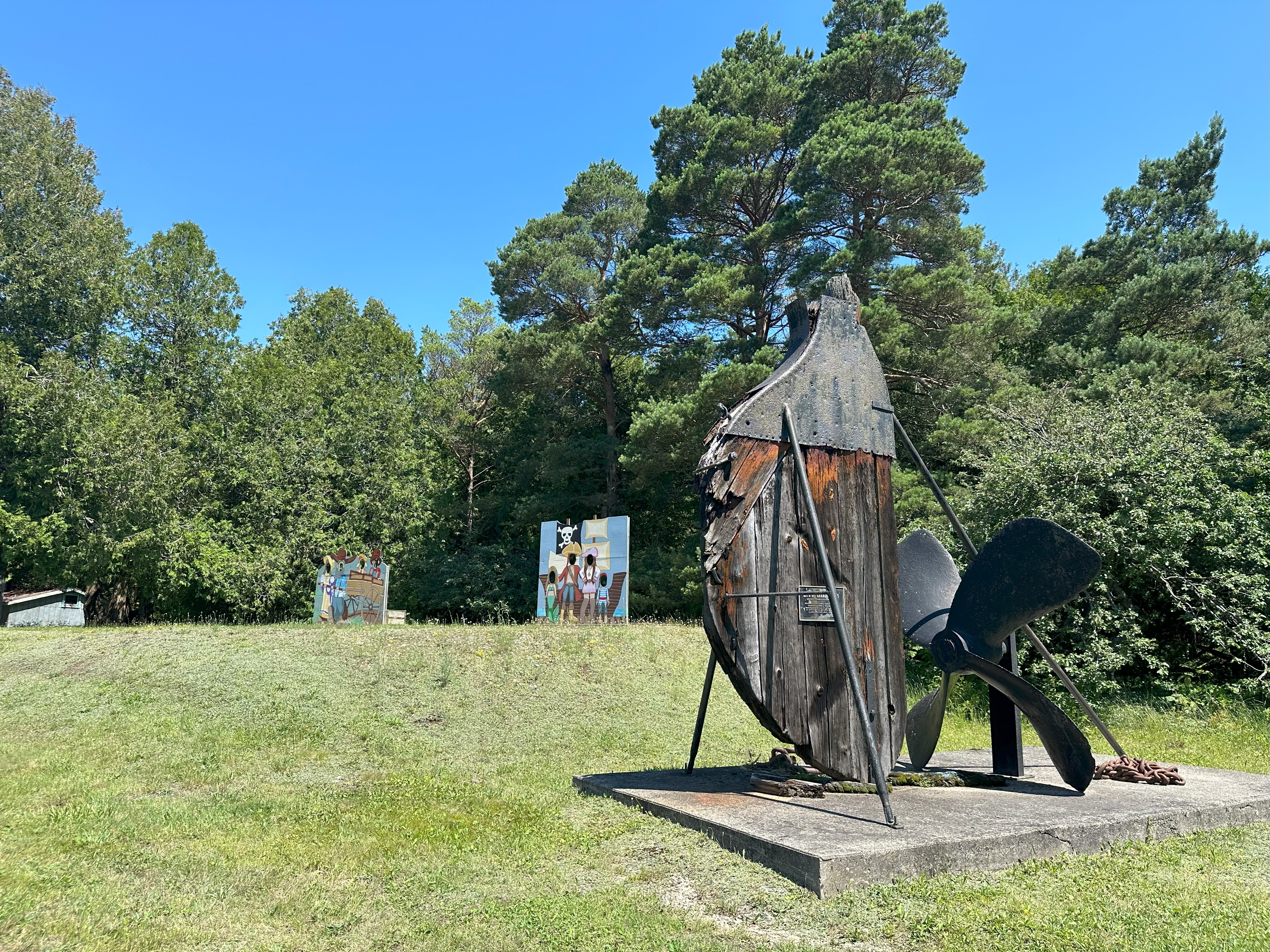 Wide view of a ship's rudder on display, outdoors in a grassy area with a rudder installation and surrounding trees.