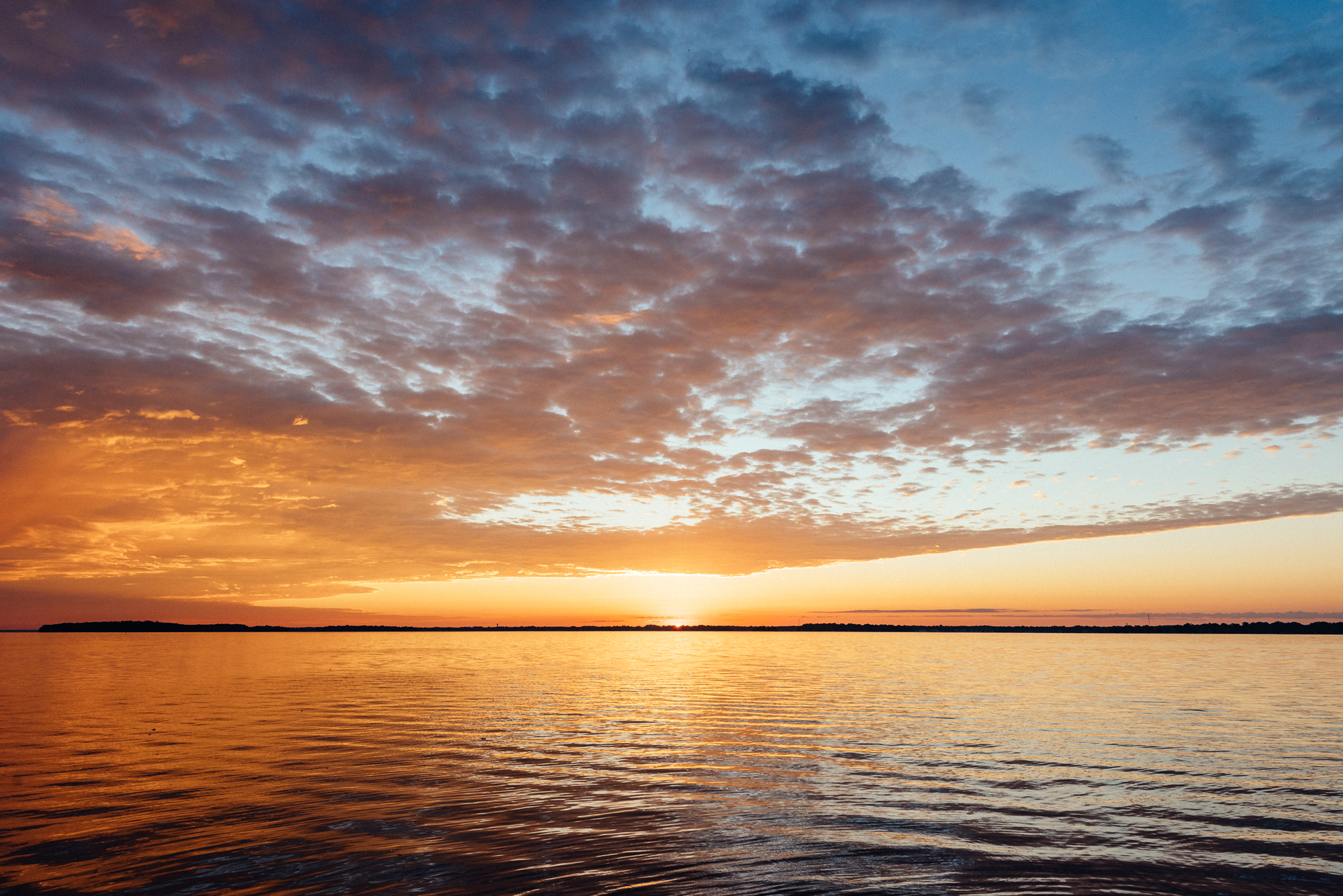 The sun sets over Lake Couchiching, casting warm orange light across calm water while scattered clouds glow in shades of gold, pink, and blue along the horizon.