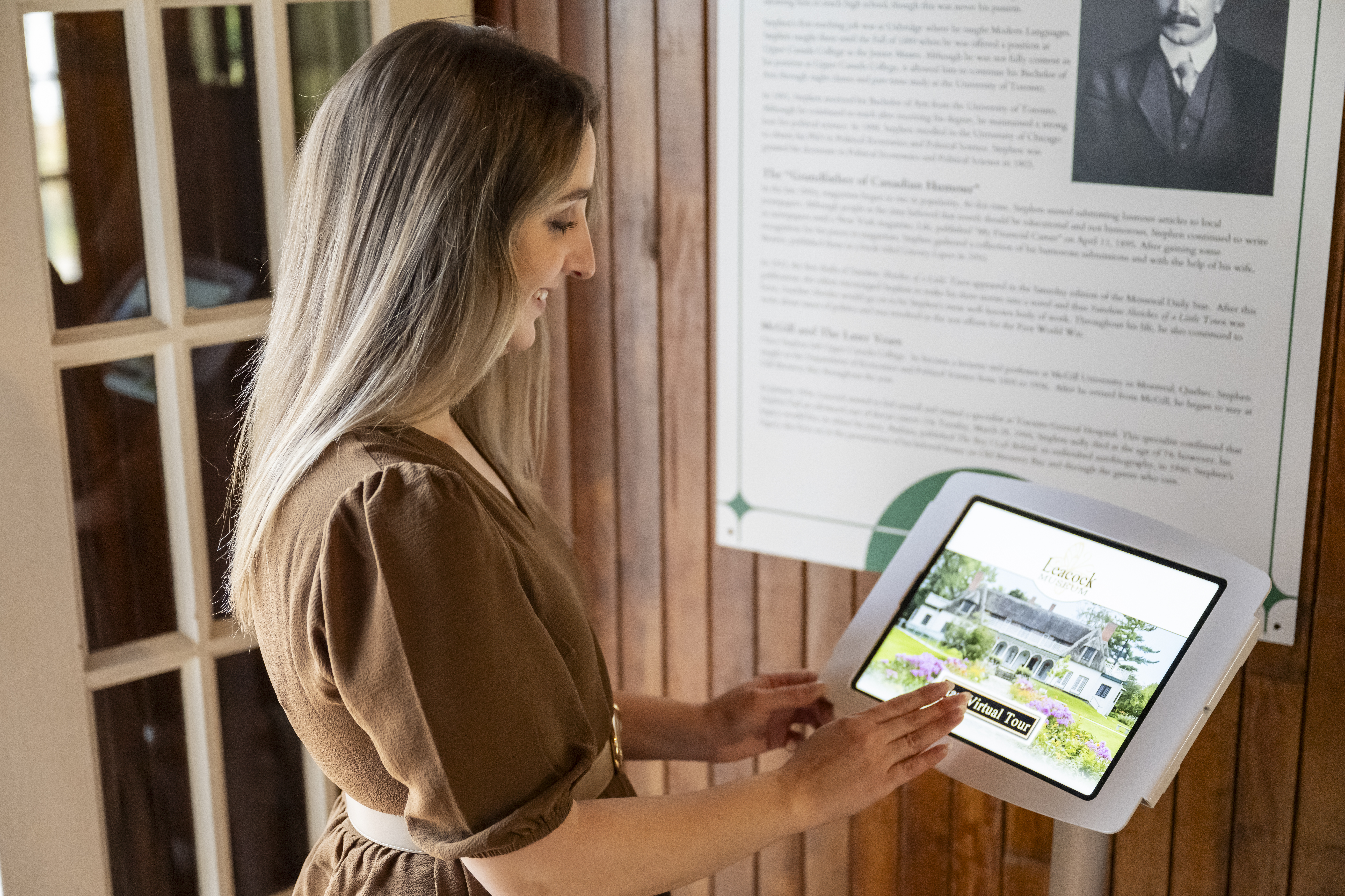 A woman uses a touchscreen display at an Orillia heritage site, exploring information about a historic property inside a wood-paneled interior.