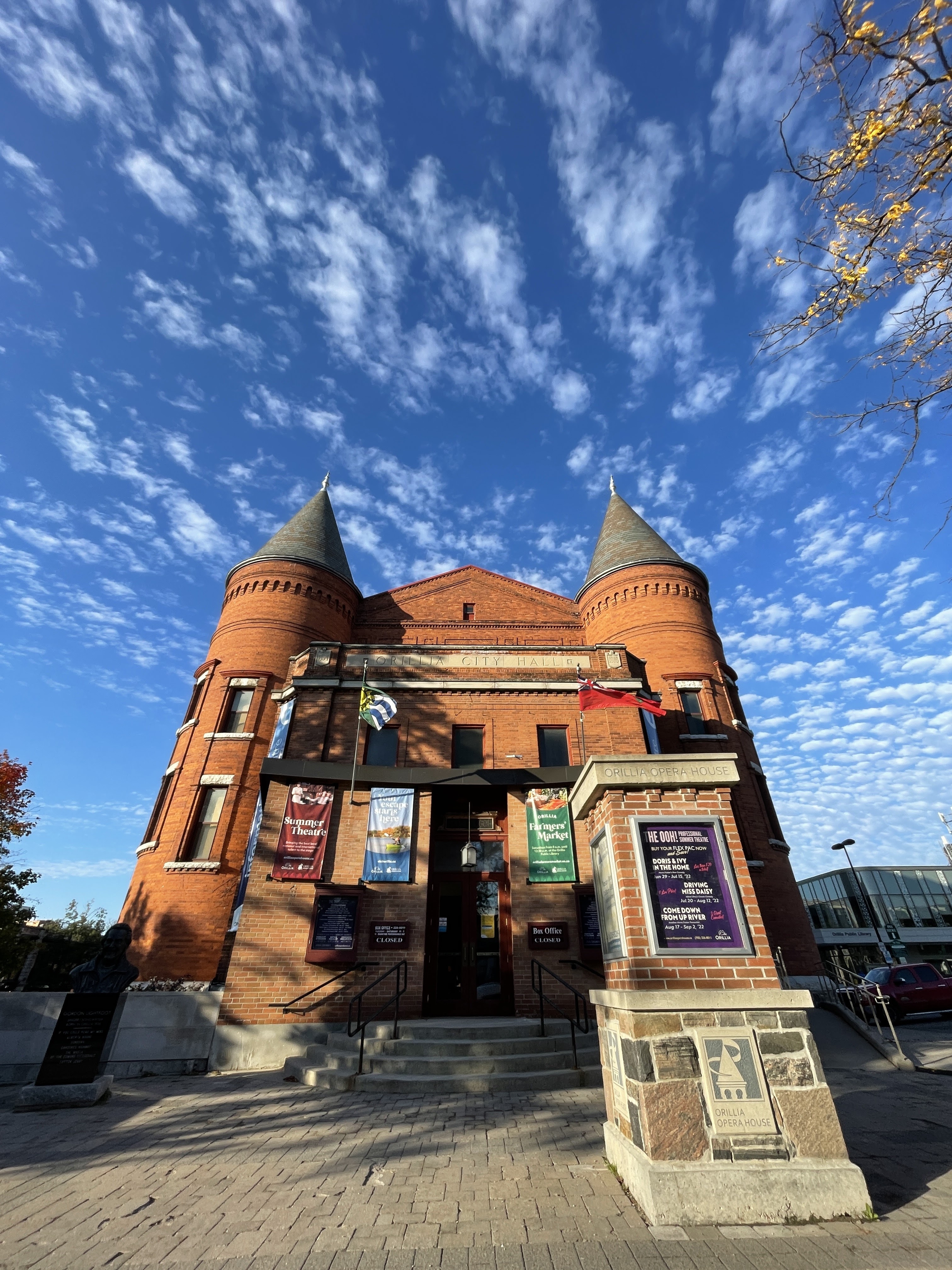 Front view of the historic Orillia Opera House with its red brick façade, twin turrets, and theatre banners under a bright blue sky with scattered clouds.