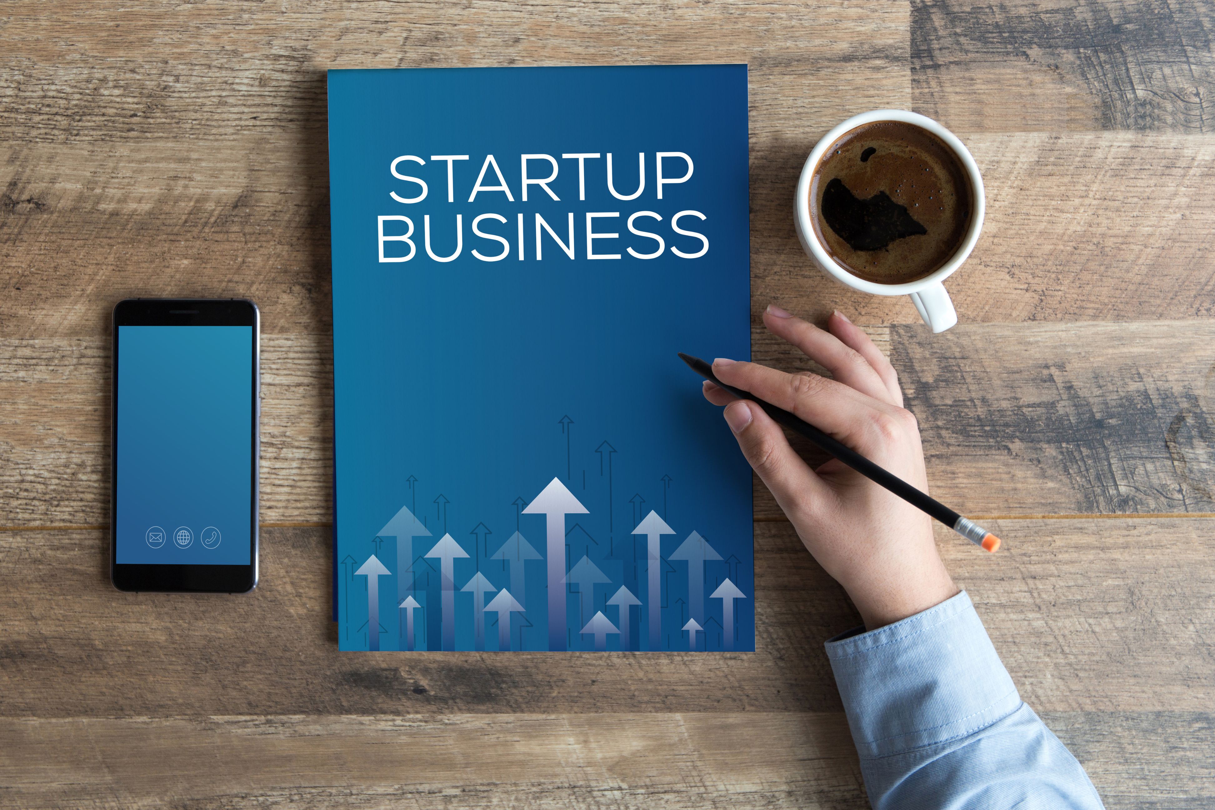 a cell phone, notebook, with a title "Startup Business", and a coffee cup on a table, with a hand holding a pencil resting on the book