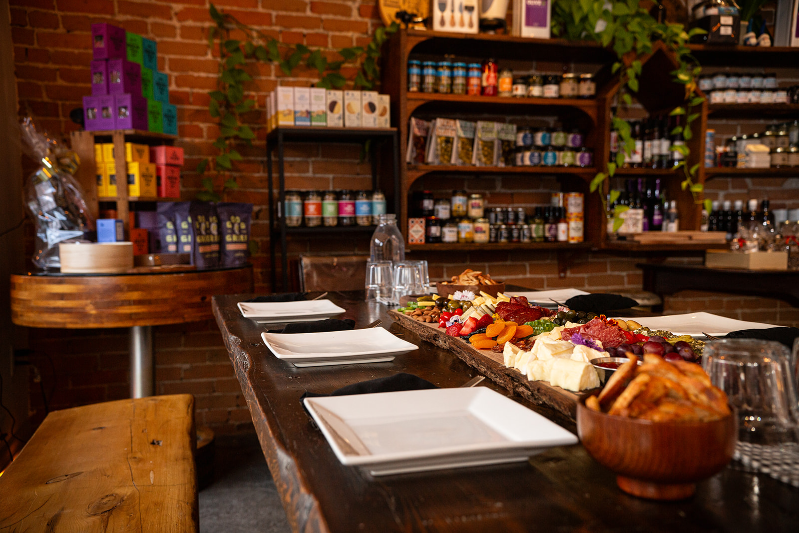 A wooden harvest table at The Milk Maid set with sharing plates and an abundant, delicious-looking charcuterie board with shelves of pickles, jams, and other grocery condiments stocked in the background.