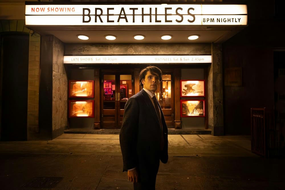 At night, a man stands out side a cinema under its marquee that reads "Now Showing: Breathless 8pm Nightly"