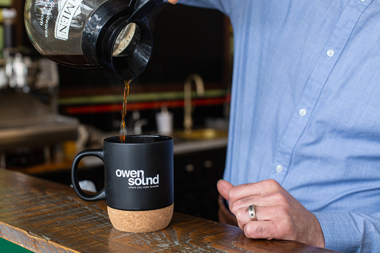 A person in a blue button-up shirt pours coffee into a black mug on the wooden counter of a cafe. The mug is printed with a variant of the Owen Sound logo, reading "Where You Want to Work."