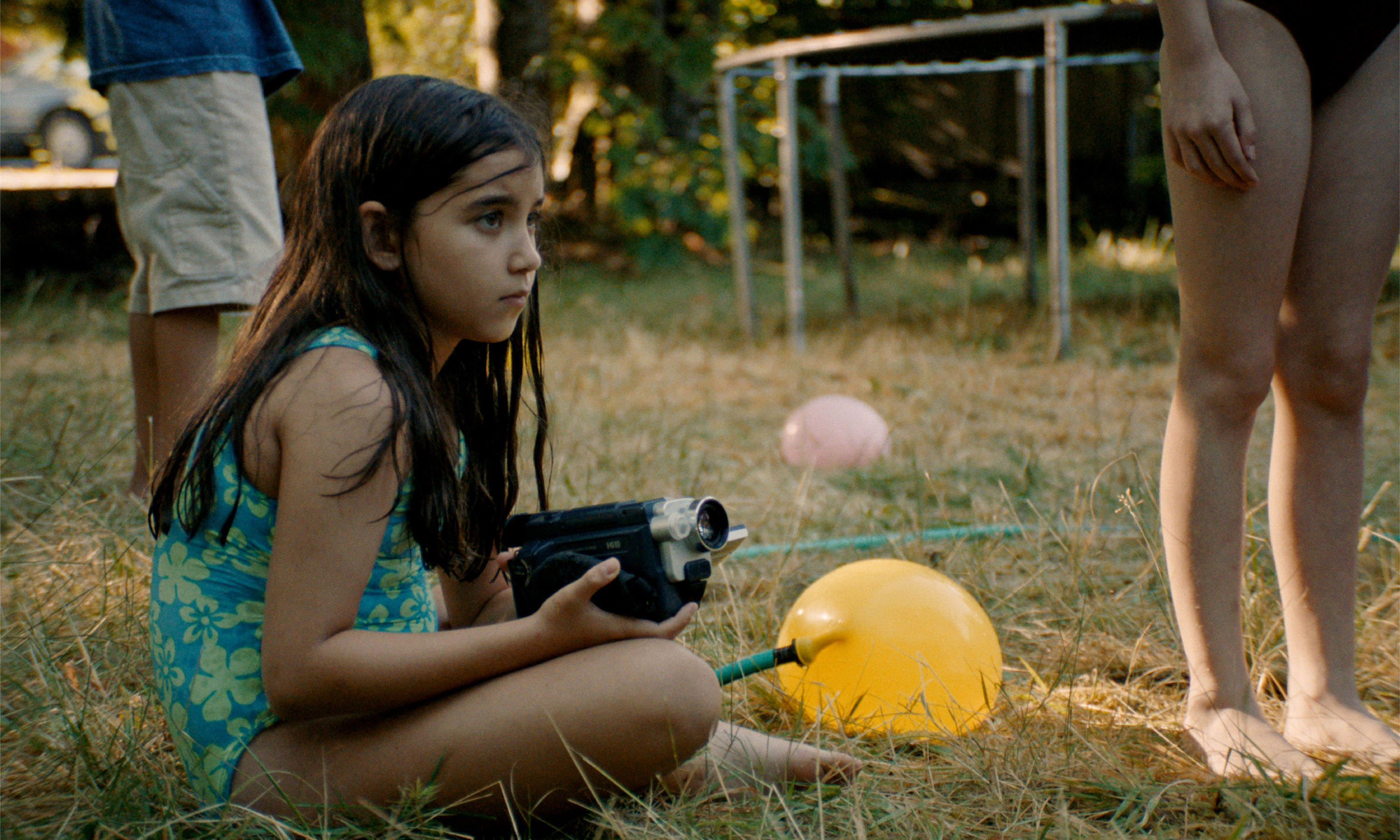 a young girl in a swim suit sits on the grass with water balloons and a video camera
