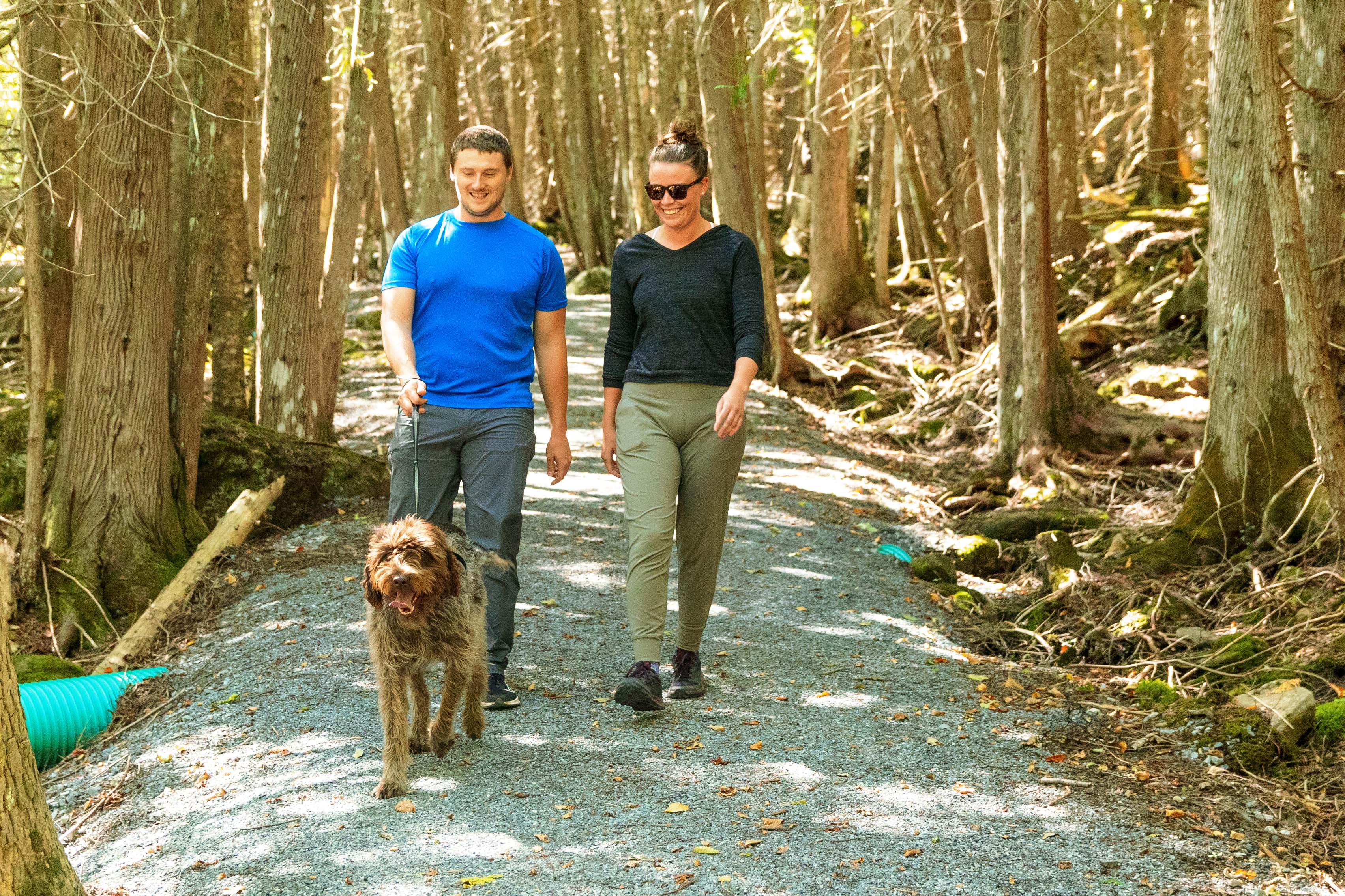 Two people walking dog on gravel trail.